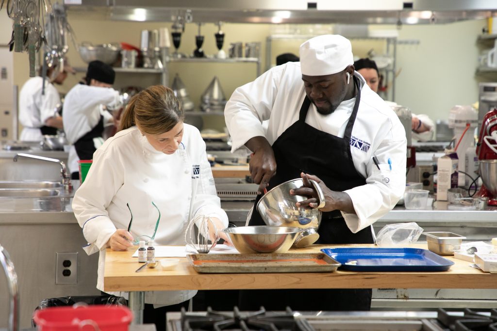 A chef instructs a male student chef as he mixes a batter in a silver bowl
