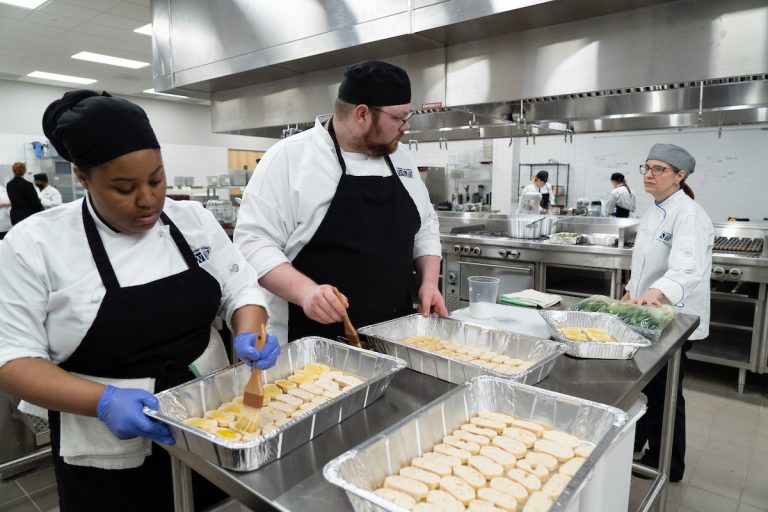 A chef instructs two student chefs as they brush butter on pans of bread.