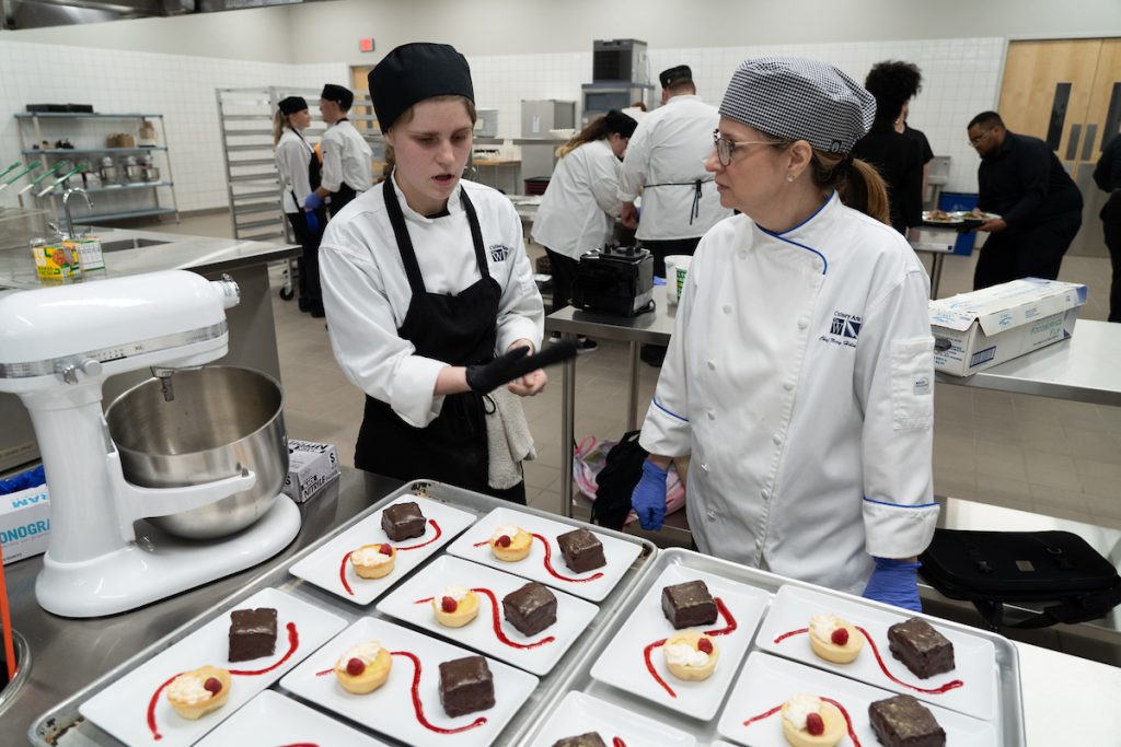 A chef instructs a student chef as they stand in front of plated desserts.