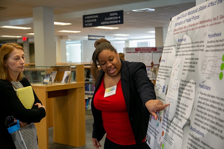 Female, African American undergraduate researcher presenting a biology poster to a faculty evaluator