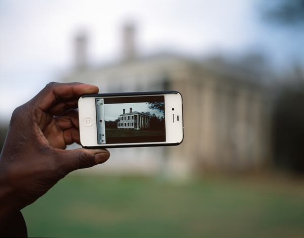 photo of hand holding iphone. historic photo of house on screen. modern house in background.
