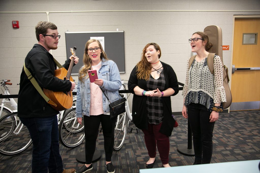 A man plays guitar and three women sing in a hallway
