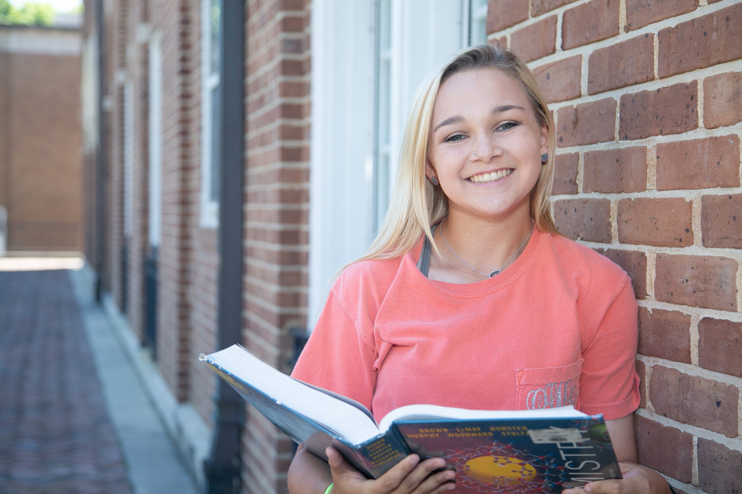 Woman with textbook leans against a brick wall