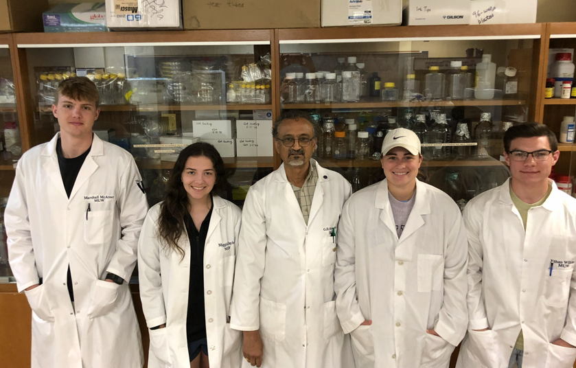 Dr. Heda and four undergraduate research students standing by a glassware cabinet.