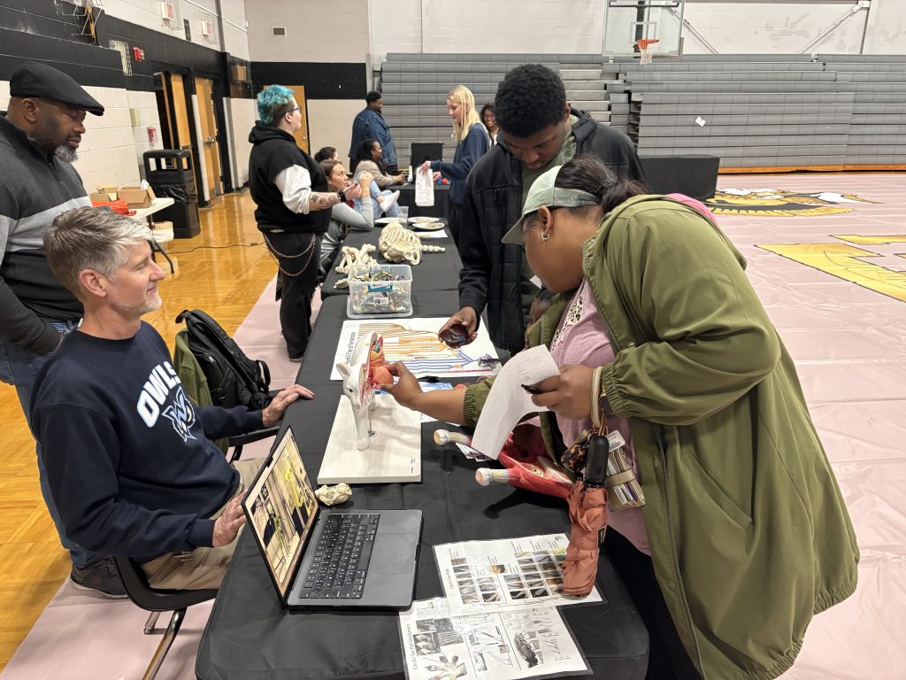 MUW faculty and students on one side of a long table, with individuals on the other side viewing displays and activities.