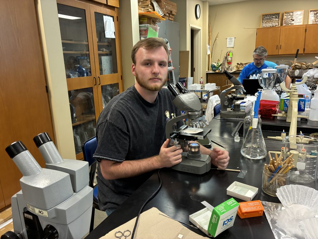 Jack Thompson at a microscope with Dr. Dodson in the background at his computer.