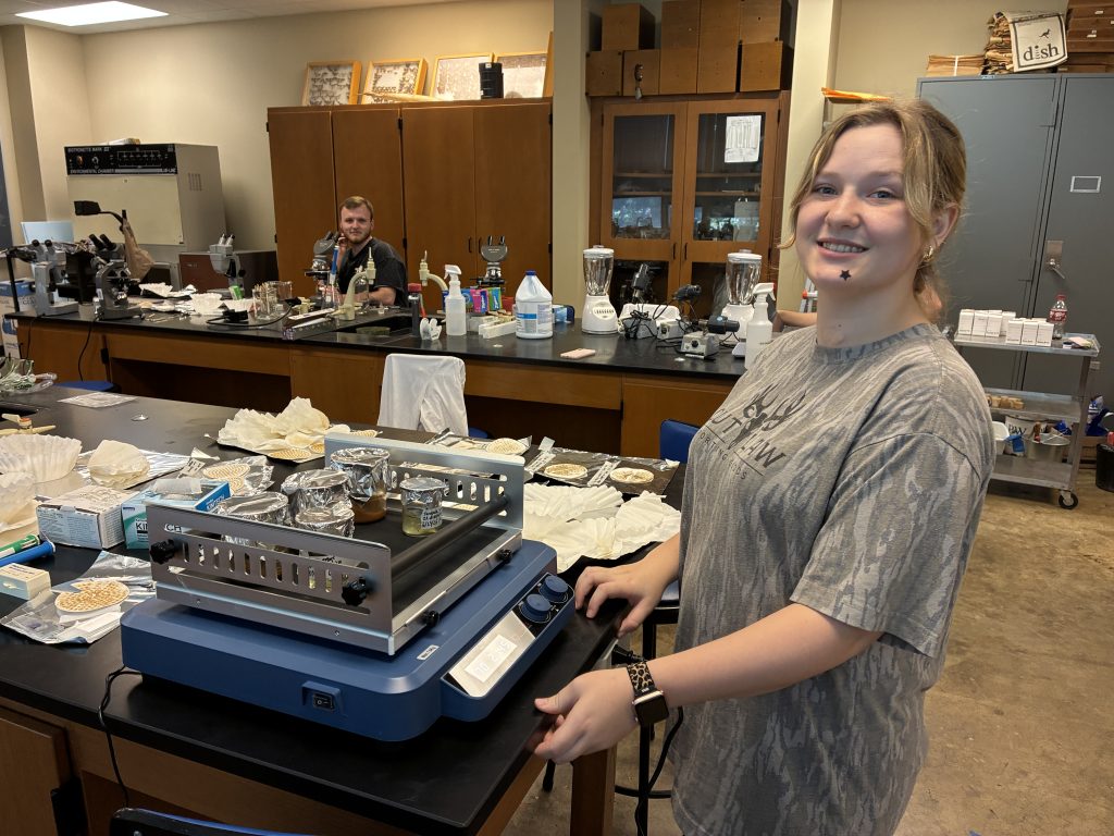 Savannah Voigt at a shaking incubator in the lab, with Jack Thompson in the background at a microscope.