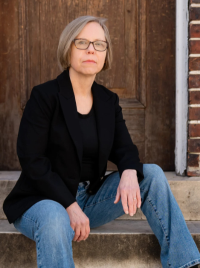 Blond woman with blond bobbed hair and black glasses, wearing black shirt and jacket in front of brown wooden door