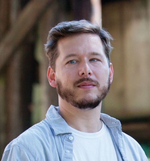 Photo of white man with short brown hair and beard, wearing open light blue shirt and white t-shirt