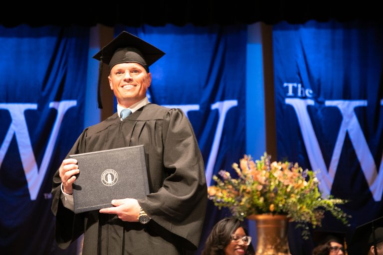 Man in cap and gown holds a degree on stage during commencement
