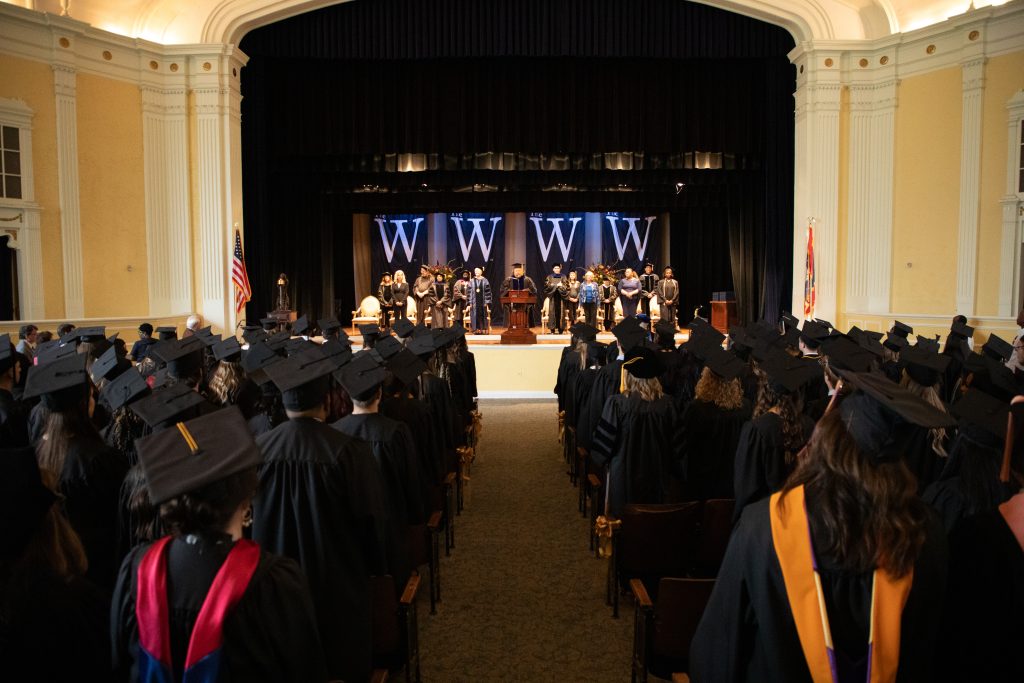 A photo of graduates in regalia facing the stage with MUW banners displayed behind the podium where the speaker addresses the crowd.