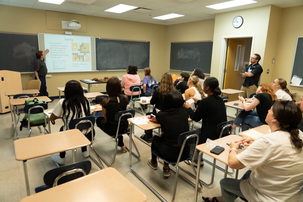 A classroom full of students look on as Dr. Hanes points to a projector screen.
