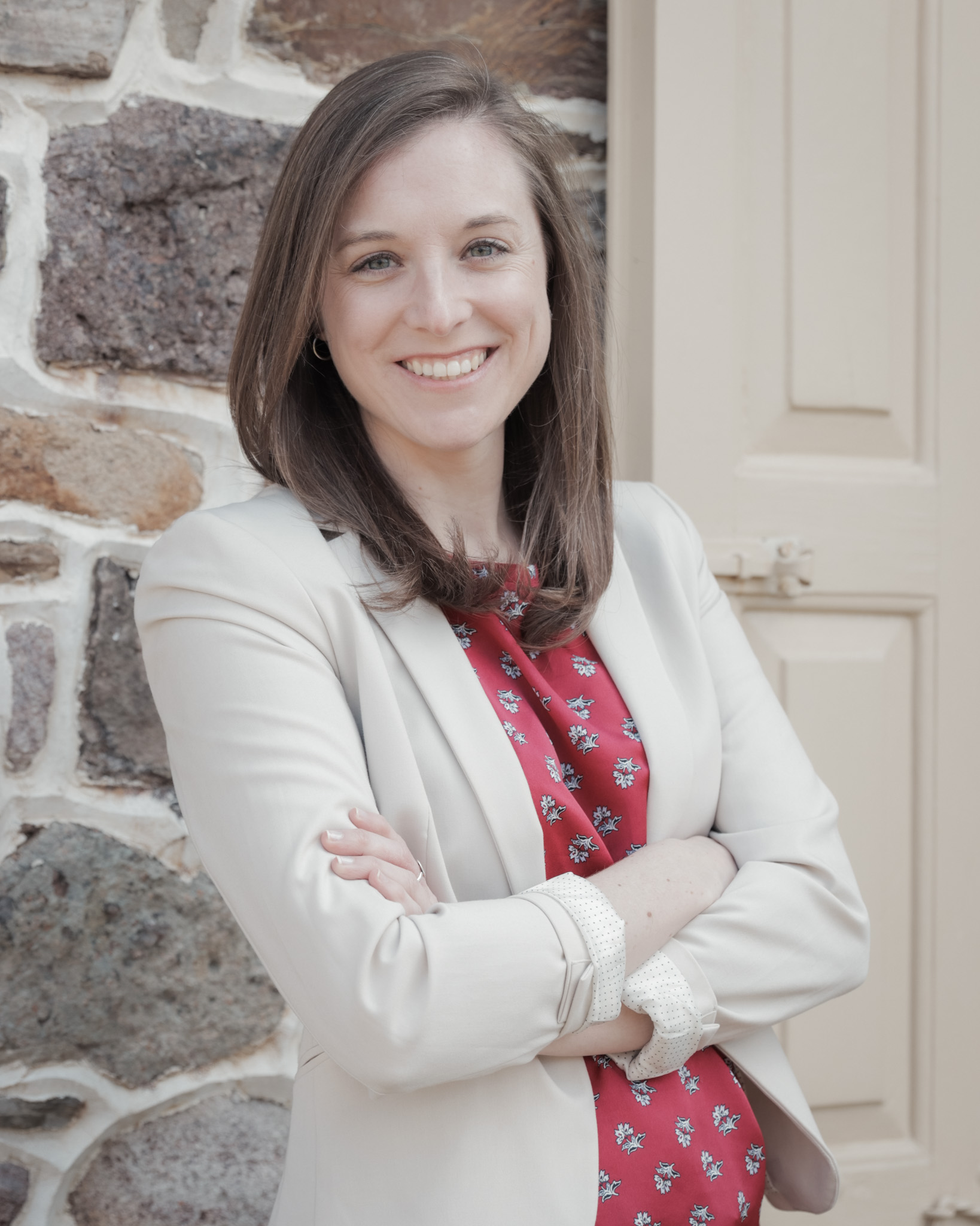 Headshot of Dr. Jacqueline Beatty. She stands smiling and leaning against a stone wall, arms crossed. She wears a cream blazer and red blouse.