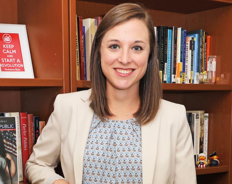 Headshot of Dr. Jacqueline Beatty. She stands smiling in front of a bookshelf, wearing a cream blazer and blue blouse. On the shelf behind her, a framed graphic reads "Keep Calm and Start a Revolution."
