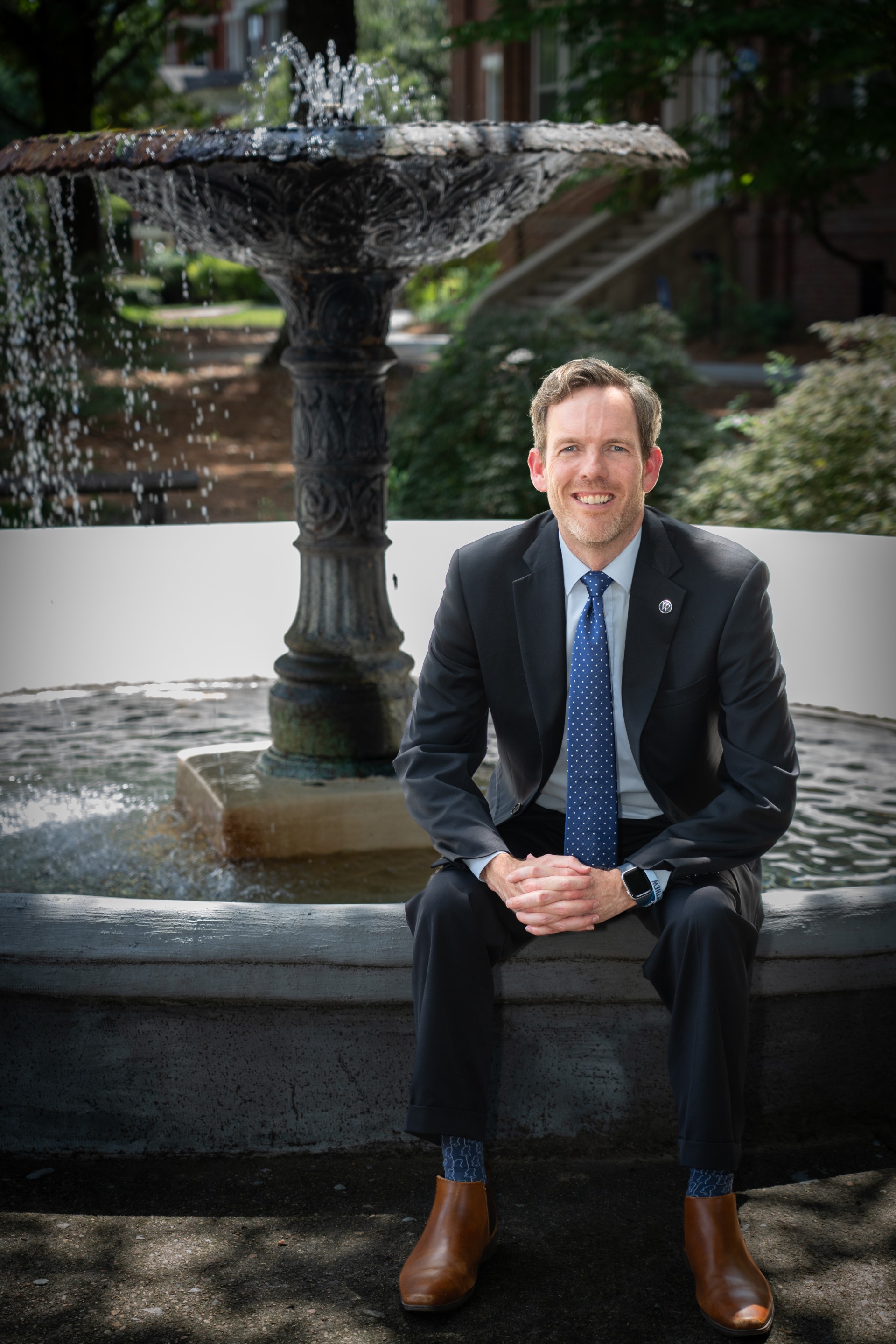 Interim President Scott Tollison, Ph.D. sits on the edge of a water fountain outdoors in official photograph