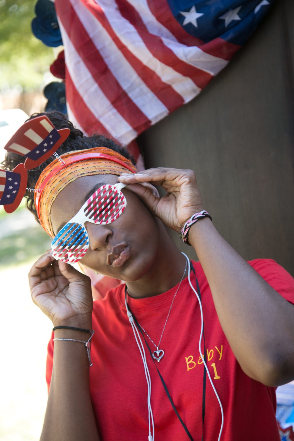 A young woman purses her lips, smiles slightly, and tilts her head as she poses in American flag-themed sunglasses and with Uncle Sam-style hats bobbing from a headband.