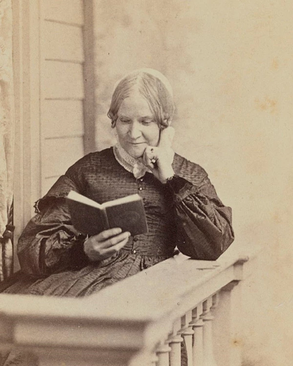 An aged black and white photograph shows Lydia Maria Child sitting on a porch, reading from a book.